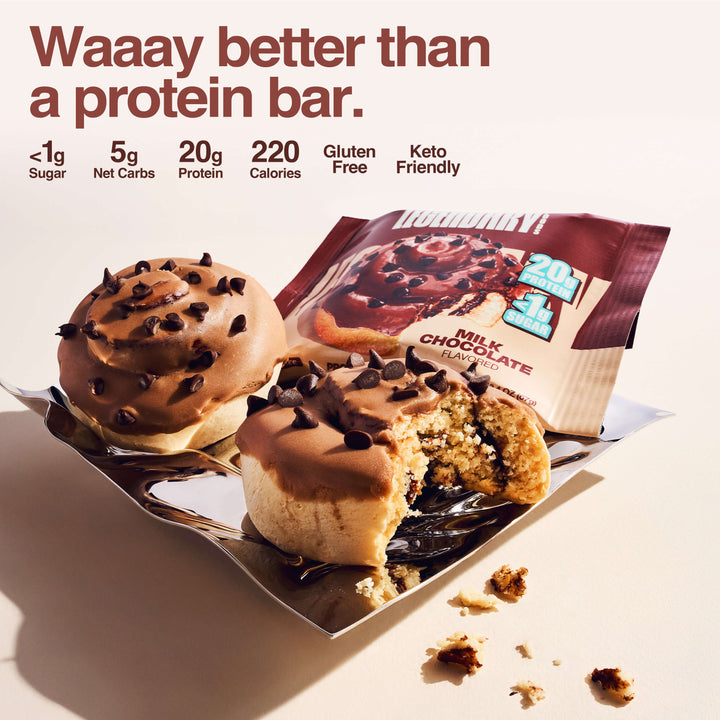 Two Milk Chocolate Protein Sweet Rolls are shown on a reflective tray—one whole and one bitten into—revealing a soft, fluffy inside. Behind them is the product packaging, which highlights "20g Protein" and "<1g Sugar". Bold text reads: “Waaay better than a protein bar.” Nutritional callouts include: <1g Sugar, 5g Net Carbs, 20g Protein, 220 Calories, Gluten Free, Keto Friendly.