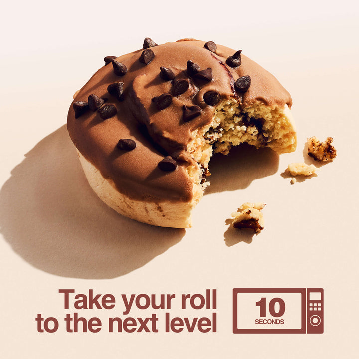 A single Milk Chocolate Protein Sweet Roll with a bite taken out sits on a cream-colored background. Crumbs are scattered nearby. Text below the roll reads: “Take your roll to the next level,” with a microwave icon showing “10 seconds,” suggesting to warm it for enhanced flavor.
