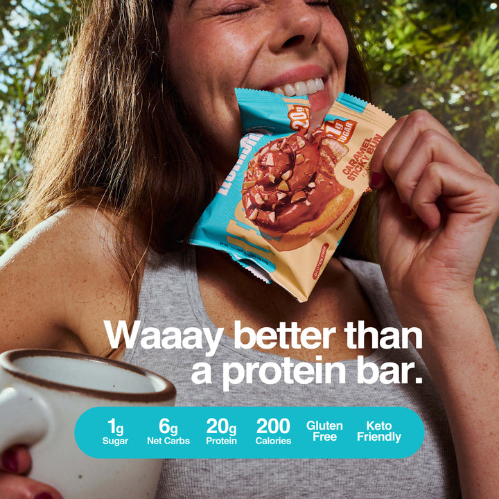 Woman smiling and holding a caramel sticky bun protein snack and coffee, with product nutrition info and text reading “Waaay better than a protein bar.”