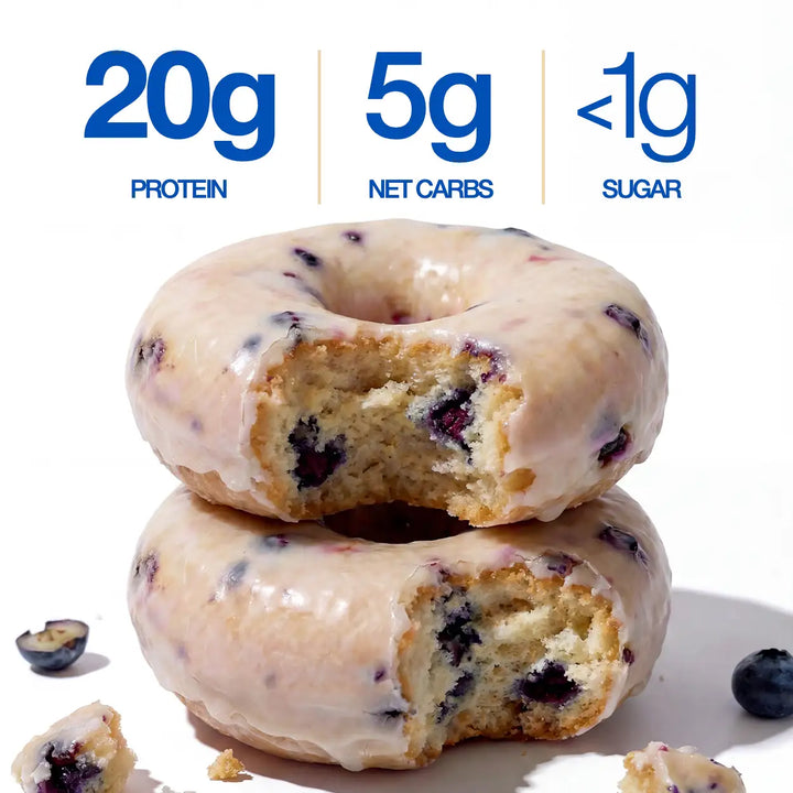 Two glazed blueberry donuts stacked on a white background, with text above reading 20g protein, 5g net carbs, and <1g sugar. Blueberries and crumbs are scattered around.