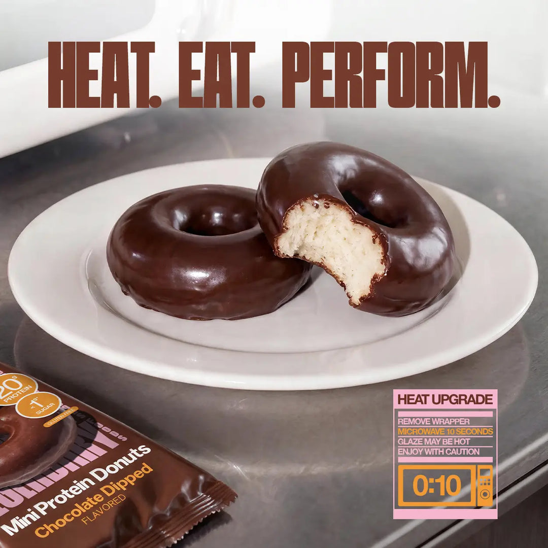 Two chocolate-dipped mini protein donuts on a white plate, with one partially bitten to show the interior. Large text at the top reads 'HEAT. EAT. PERFORM' with optional instructions to microwave the donuts for 10 seconds.