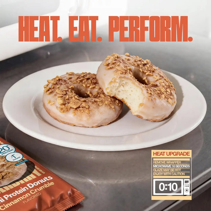 Two Cinnamon Crumble Mini Protein Donuts on a white plate, one partially eaten, with the headline "Heat. Eat. Perform." and instructions to microwave unwrapped donuts for 10 seconds.