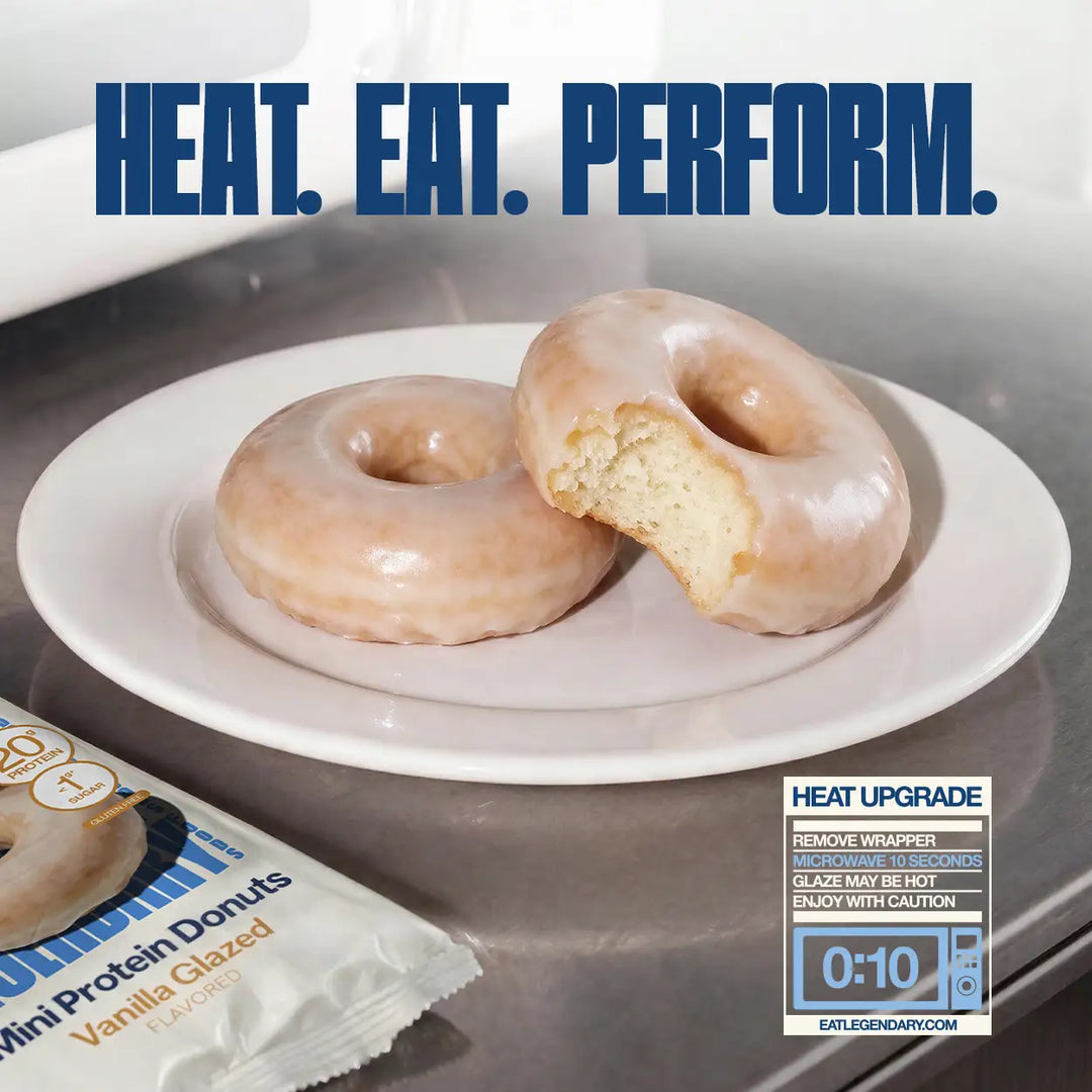 Two Vanilla Glazed Mini Protein Donuts on a white plate, one with a bite removed. Heating instructions recommend microwaving unwrapped donuts for 10 seconds.