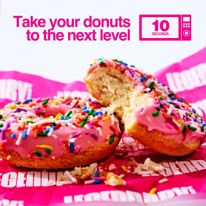 Two pink-frosted donuts with rainbow sprinkles, one partially eaten, sit on pink Legendary Foods paper. Text says “Take your donuts to the next level – 10 seconds” with a microwave icon.
